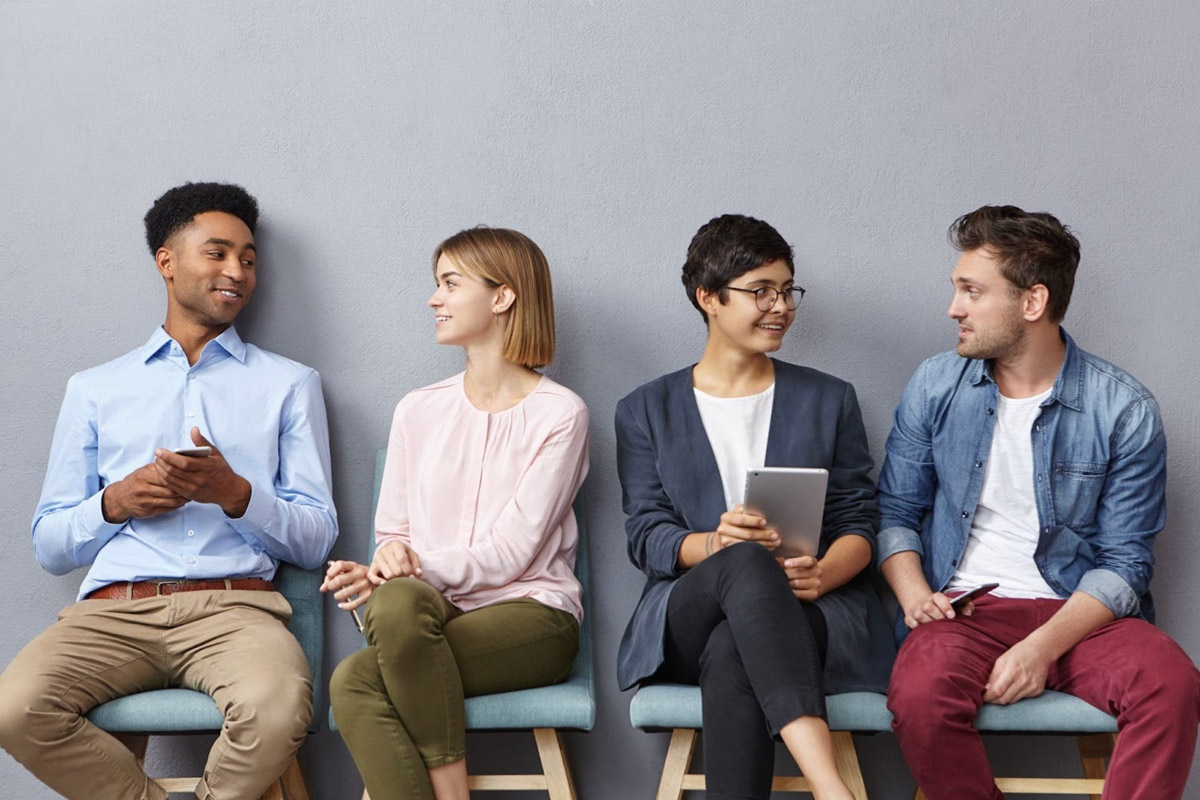 Four diverse professionals seated in a row, engaging in conversation and holding electronic devices, symbolising collaboration and the role of a recruitment partner in connecting talent.