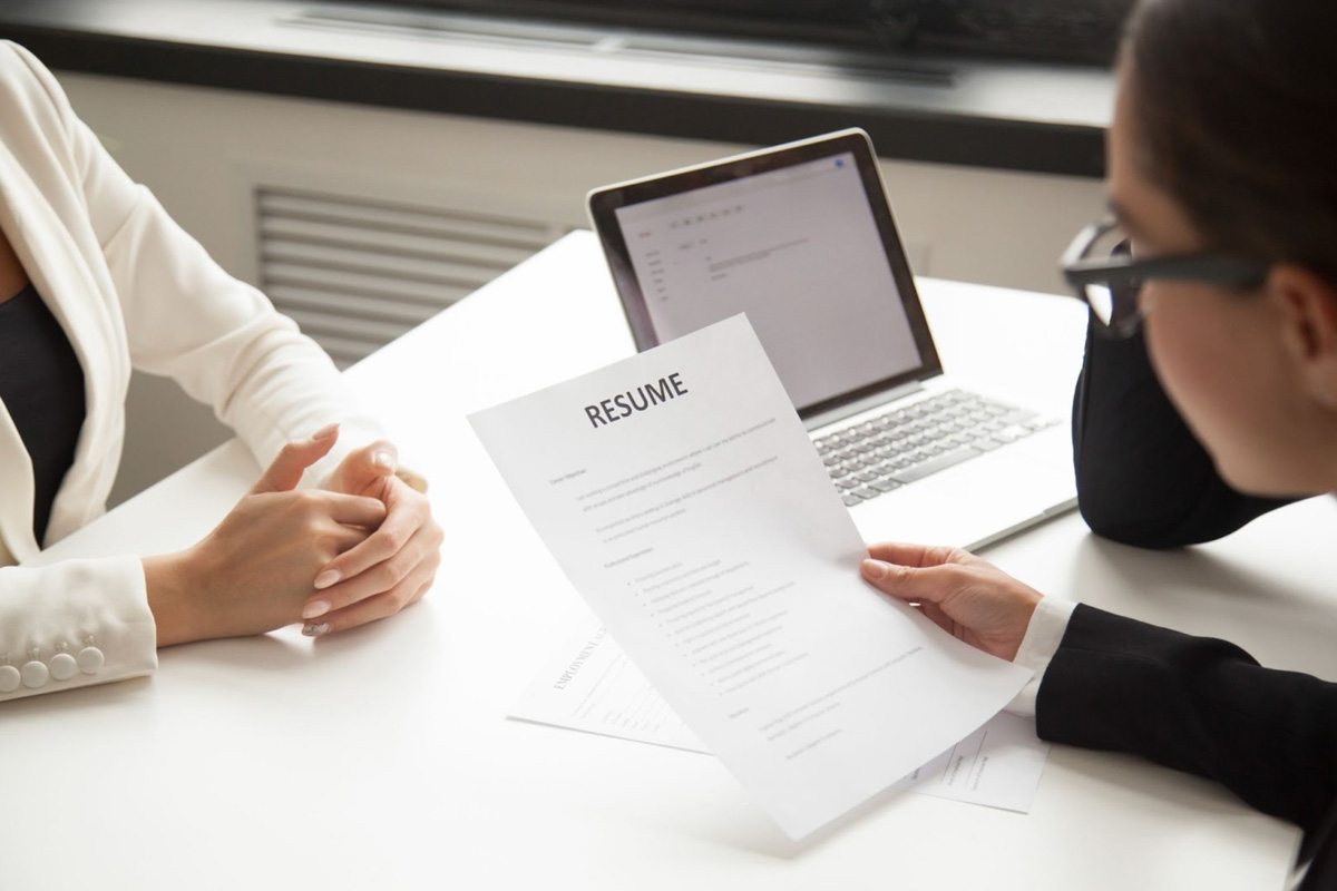Professional job interview setting with a recruitment partner reviewing a candidate's resume at a white table, accompanied by a laptop in a bright office environment.