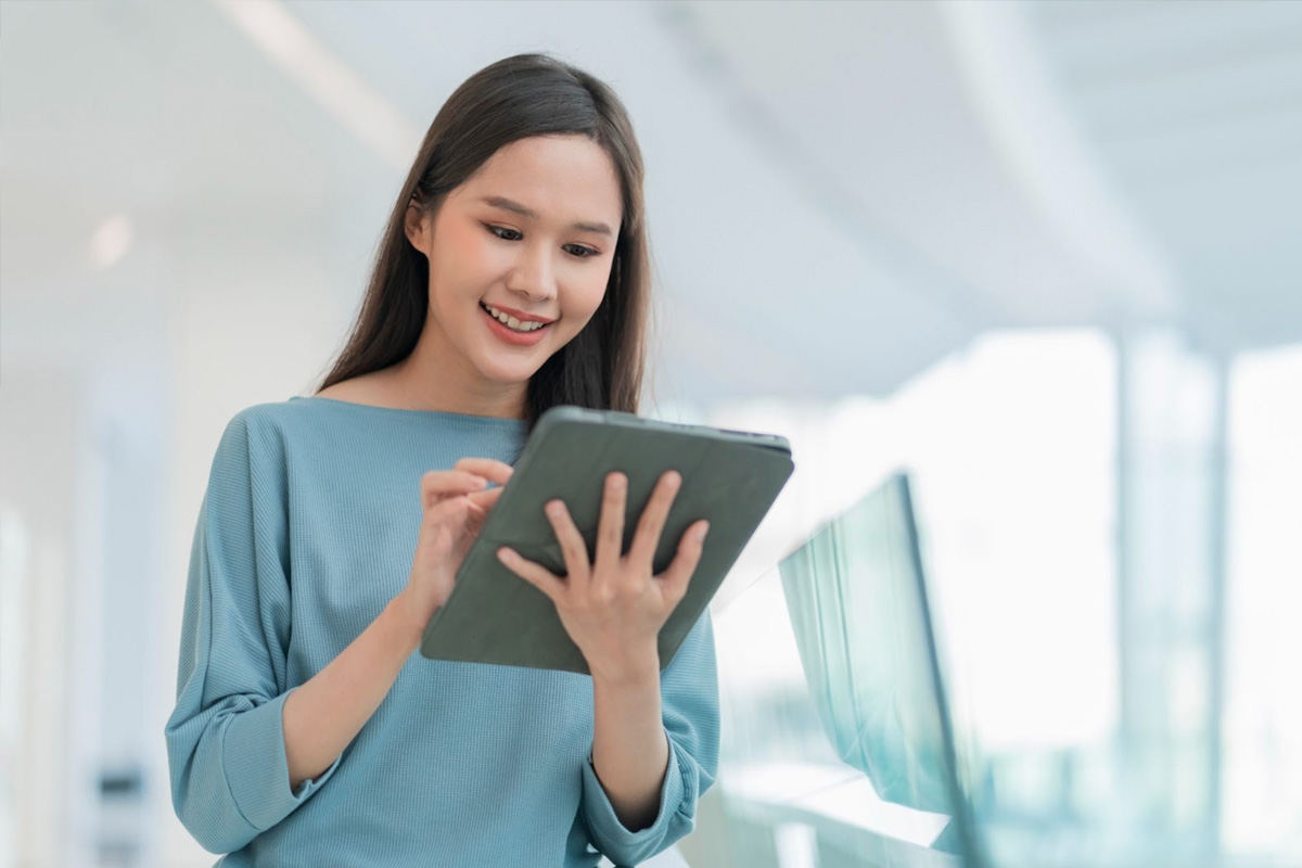 Smiling young Asian woman using a digital tablet in a bright, modern indoor setting, used for recruitment